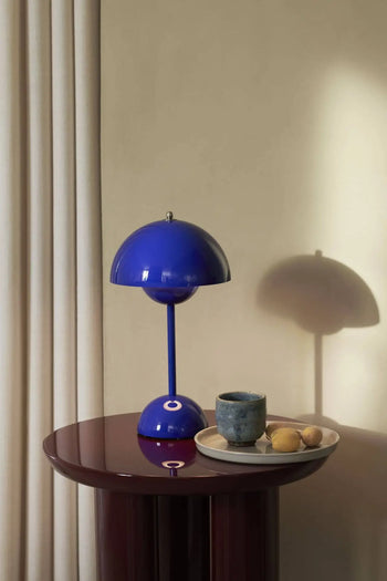 An &Tradition Flowerpot VP9 Portable Table Lamp sits on a glossy maroon side table beside a ceramic cup and two lemons on a plate, with cream-colored curtains and a beige wall in the background.