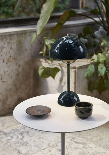 A round white table displays an &Tradition Flowerpot VP9 Portable Table Lamp in dark green, a black ceramic bowl, and a black cup. A USB charging cable is beside the lamp, with large leafy plants and a stone wall in the background.
