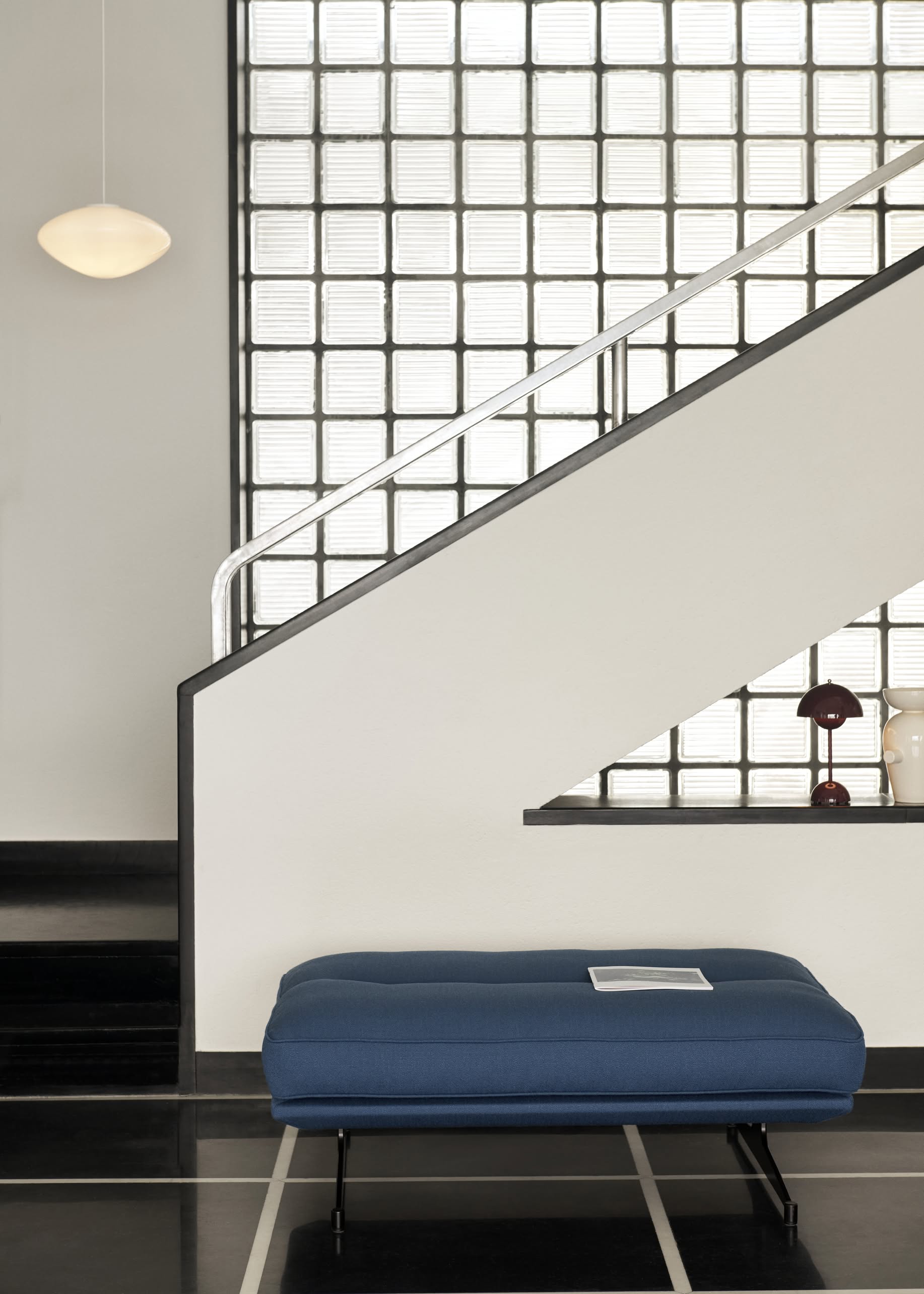 A minimalist interior features a staircase with glass-block walls, metallic handrail, blue bench with a paper on top, and decorative accents like the &Tradition Flowerpot VP9 Portable Table Lamp displayed on a shelf beneath the stairs.
