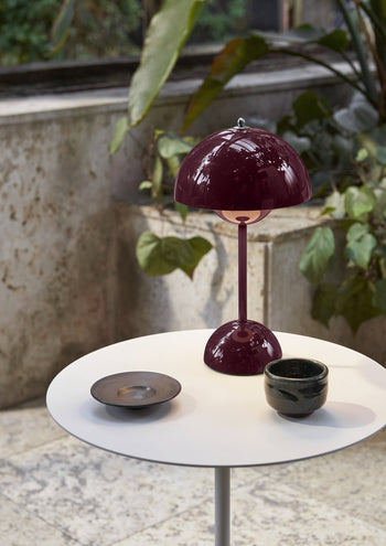 A small white round table displays an &Tradition Flowerpot VP9 Portable Table Lamp in dark red, a ceramic cup, and a dark plate with a USB charging cable nearby. Green plants and a textured stone wall form the backdrop.