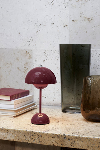 The &Tradition Flowerpot VP9 Portable Table Lamp by &Tradition, featuring a dome shade in burgundy, sits on stone beside stacked books and glass vases. This portable lamp comes with a USB charging cable and is set against a pale textured stone wall.