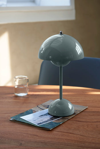 An &Tradition Flowerpot VP9 Portable Table Lamp sits on magazines atop a wooden desk beside a glass of water, while sunlight pours through the window, creating gentle shadows.