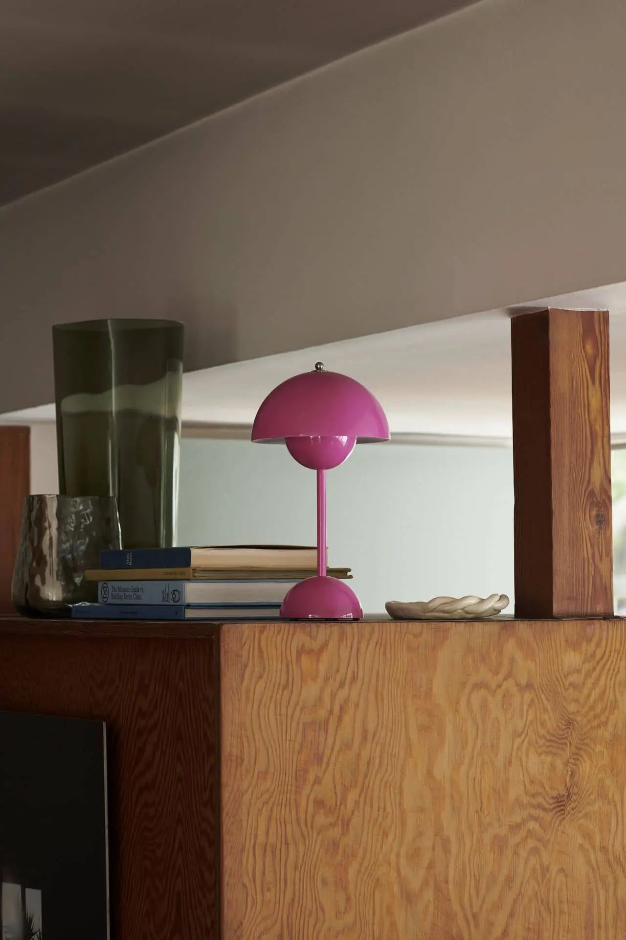 The &Tradition Flowerpot VP9 Portable Table Lamp adds a stylish touch on a wooden shelf beside a green glass vase, textured clear glass, books, and ceramic dish. Its minimalist setting highlights the lamp’s design and USB charging cable.