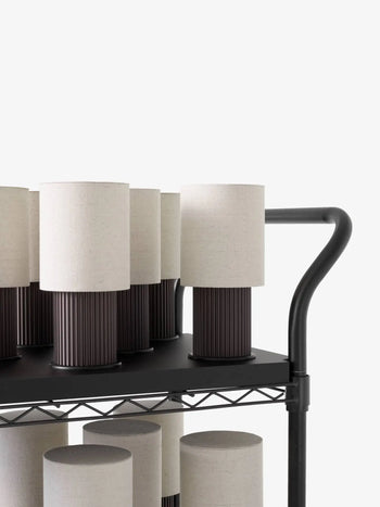 Several &Tradition Manhattan SC52 Portable Table Lamps by &Tradition, featuring cylindrical off-white fabric shades and dark ribbed bases, are arranged on a black metal cart with a wire shelf and curved handle against a white background.