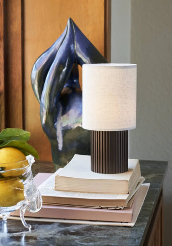 The &Tradition Manhattan SC52 Portable Table Lamp with a pleated dark base and white shade is displayed on stacked books beside a glass bowl of lemons and a modern blue ceramic sculpture on a marble surface.