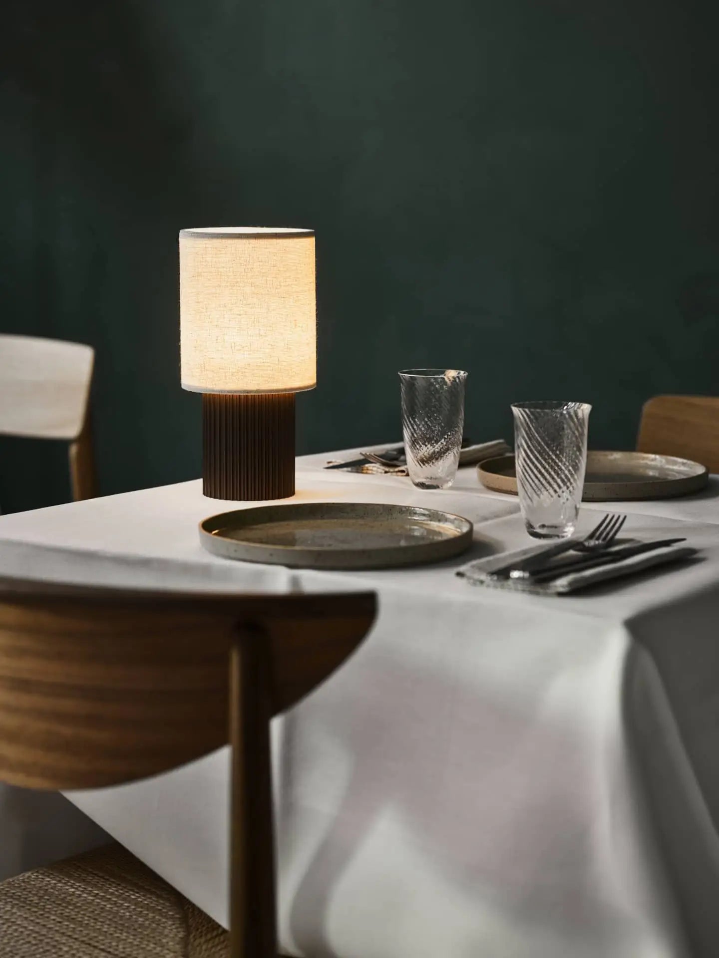 A neatly set dining table with a white tablecloth, ceramic plates, cutlery, and the &Tradition Manhattan SC52 Portable Table Lamp by &Tradition. Wooden chairs surround the table, with a dark green wall adding contrast to this industrial-inspired space.