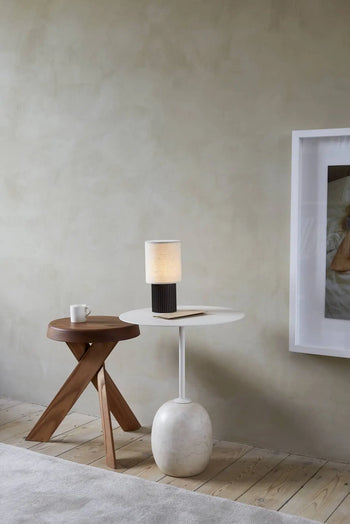 A minimalist interior includes a small wooden stool with a mug, a white round side table with an &Tradition Manhattan SC52 Portable Table Lamp and book, plus a framed photo on a beige wall. Light wood floors are partly covered by a gray rug.