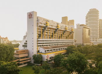 Bathed in warm sunlight, a large white and yellow building with geometric balconies rises among trees and high-rises, echoing the Golden Mile Complex—showcased in UNIT. Volume 2: Golden Mile Complex by UNIT.