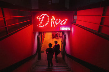 Two people descend dimly lit stairs in Golden Mile Complex, heading toward a hallway lit by bright neon Diva and TODAYS signs, creating a vibrant red atmosphere—featured in UNIT. Volume 2: Golden Mile Complex by UNIT.