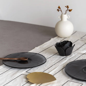 A minimalist table setting features the Urban Nature Culture Breakfast Plate Recycled Glass, a wooden spoon, gold leaf decor, and a black flower bowl; a white vase with dried flowers sits on a striped runner—ideal for sustainable living.