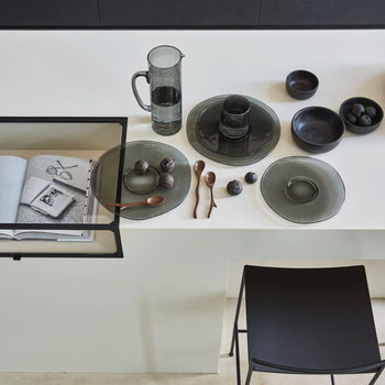 A minimalist table features Urban Nature Culture Breakfast Plate Recycled Glass, bowls of dark fruit, wooden spoons, a glass water pitcher, and an open book on a white surface beside a black chair—ideal for eco-friendly living.