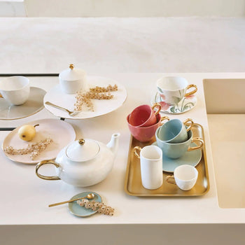 A set of elegant teacups, saucers, a teapot, and plates with gold accents are arranged on a white countertop, featuring the Urban Nature Culture Good Morning Breakfast Plate in soft pastels alongside decorative pastries on a golden tray.