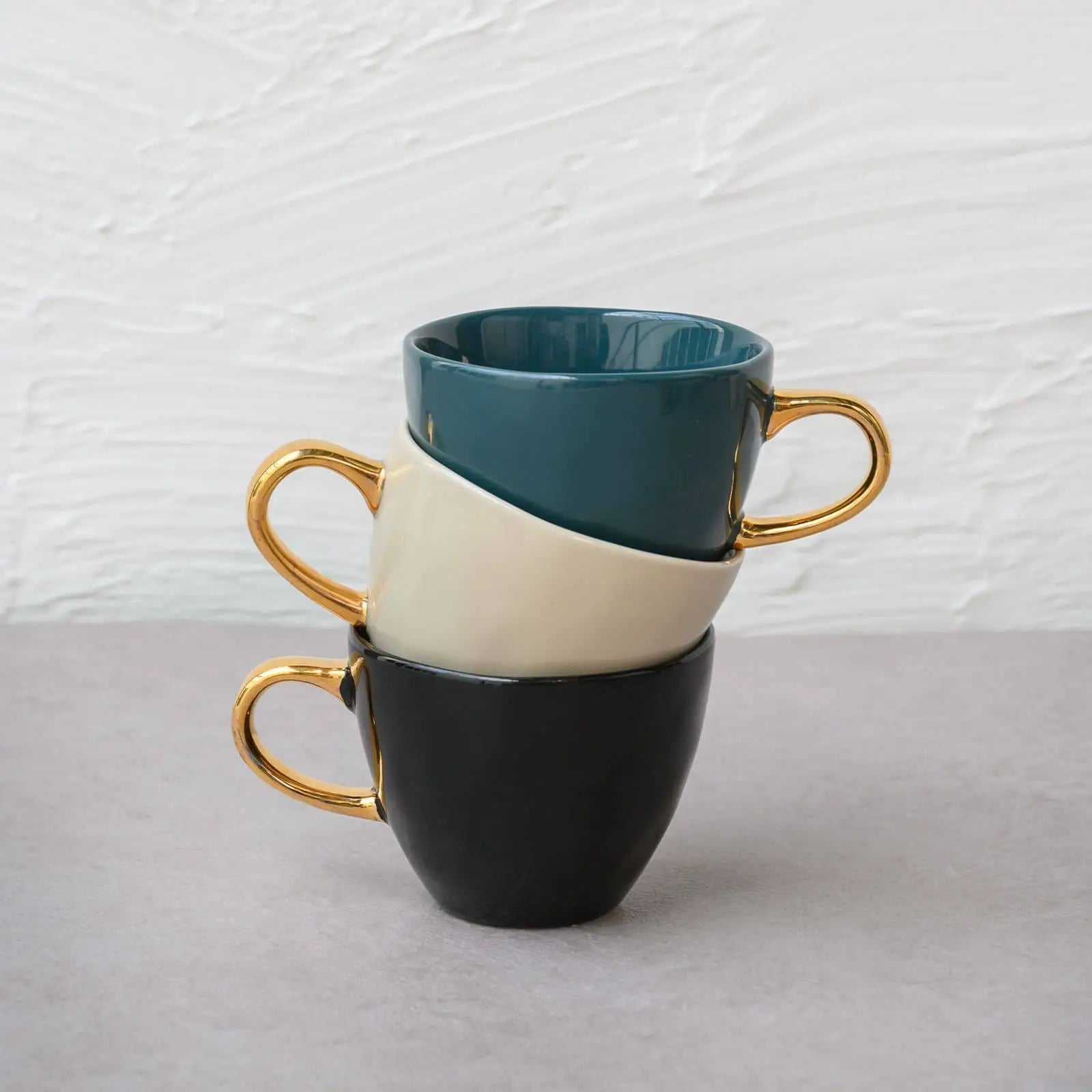 Three Urban Nature Culture Good Morning Cup Mini ceramic cups with gold handles are stacked on their sides—teal on top, cream in the middle, black on the bottom—against a light textured wall and gray surface.