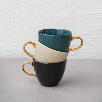 Three Urban Nature Culture Good Morning Cup Mini ceramic cups with gold handles are stacked on their sides—teal on top, cream in the middle, black on the bottom—against a light textured wall and gray surface.