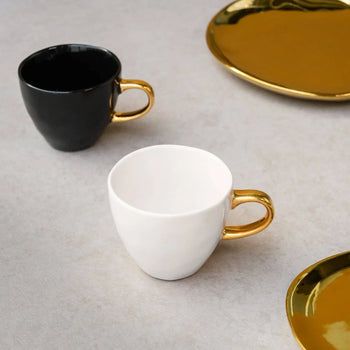 Two Urban Nature Culture Good Morning Cup Mini ceramic cups—one black, one white, each with a gold handle—rest on a light surface beside two partially visible round, gold-colored plates.