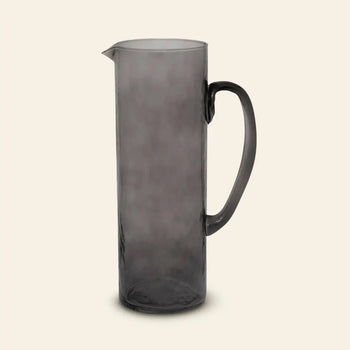 The Urban Nature Culture Jug Recycled Glass features a tall silhouette in translucent gray, with a curved handle and small pouring spout, set against a plain beige background.