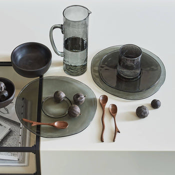 A minimal table setting with translucent gray glass plates, an Urban Nature Culture Jug Recycled Glass, a black bowl, dark plums, two wooden spoons, and a glass cup arranged on a white surface.