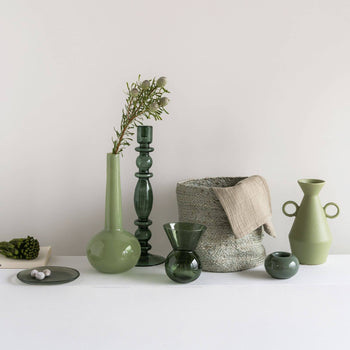 A selection of decorative green vases, bowls, and vessels in earthy tones sits on a white surface. The display includes the Urban Nature Culture Plate Recycled Glass Frost, an irregular plate by Urban Nature Culture, paired with ceramics and glassware.