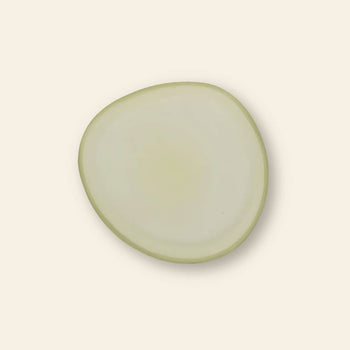 A single, irregularly shaped translucent onion slice rests on the Urban Nature Culture Plate Recycled Glass Frost by Urban Nature Culture, set against a plain, light beige background.