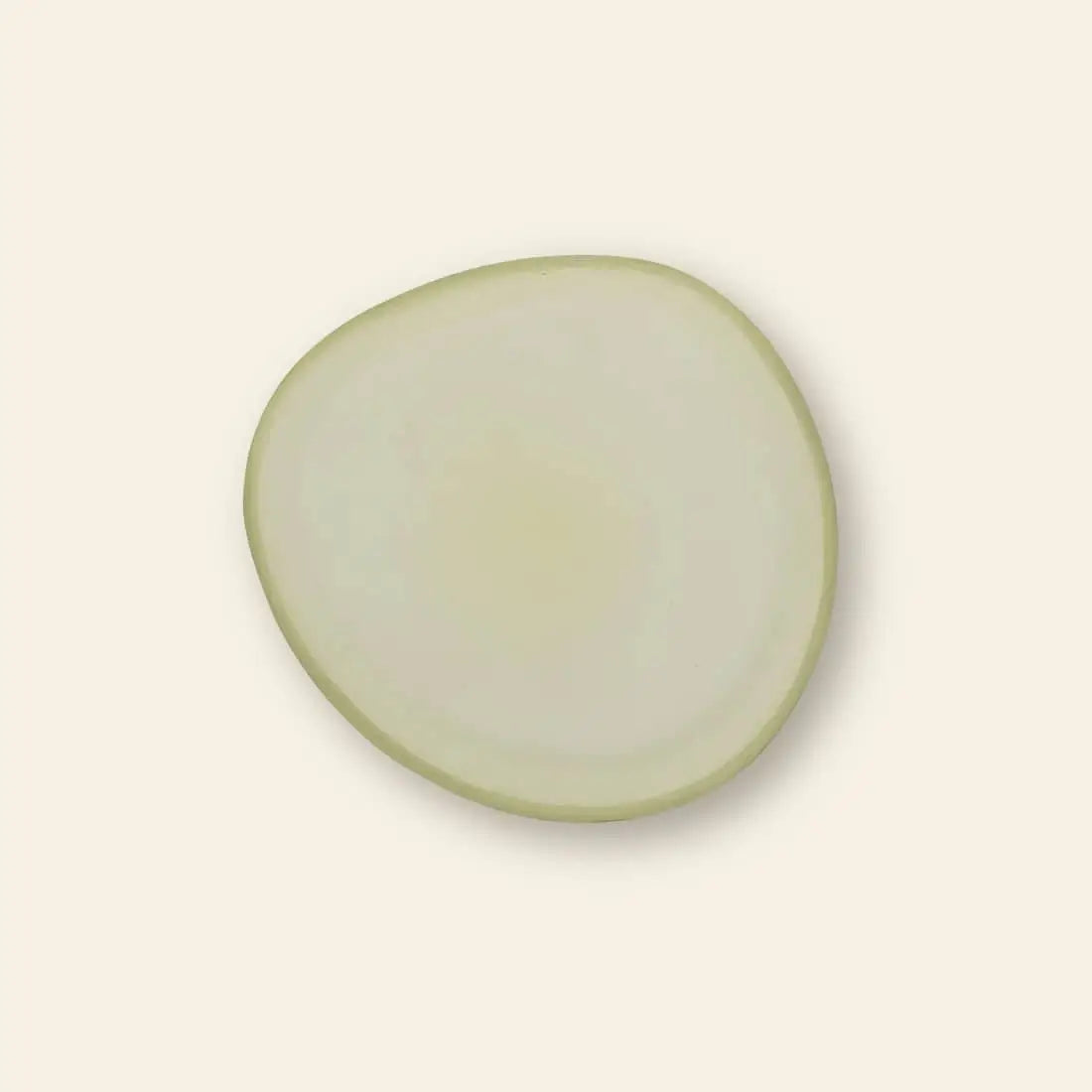 A single, thin, circular slice of onion with a pale white interior and light green edge sits on the Urban Nature Culture Plate Recycled Glass Frost by Urban Nature Culture, set against a plain off-white background.