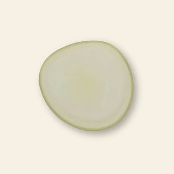 A single, thin, circular slice of onion with a pale white interior and light green edge sits on the Urban Nature Culture Plate Recycled Glass Frost by Urban Nature Culture, set against a plain off-white background.