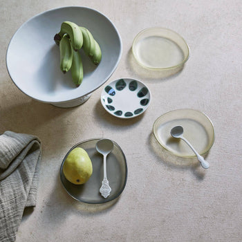 A tabletop displays Urban Nature Culture’s Plate Recycled Glass Frost, paired with bananas in a bowl, an oval plate holding a pear and spoon, a patterned dish, fabric napkin, and another spoon on a textured surface.