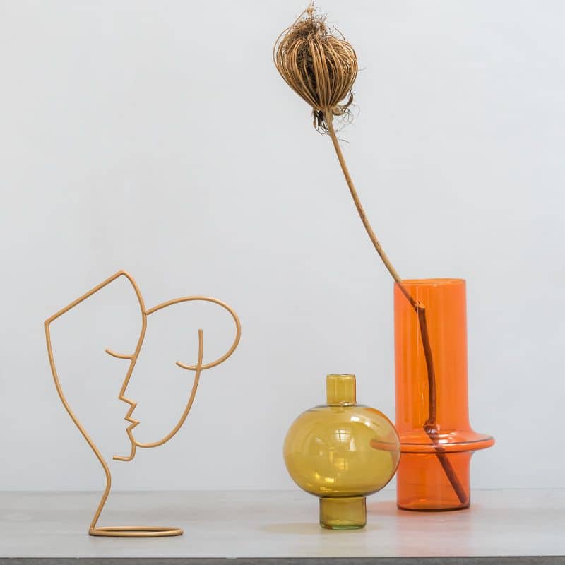 A minimalist arrangement with an abstract wire face sculpture, a small amber glass vase, and the Urban Nature Culture Vase Recycled Glass in Paprika holding a dried botanical stem against a plain background.