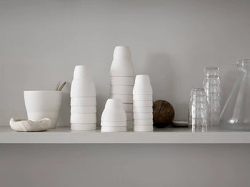 A minimalist white shelf displays stacked Vipp Vipp202 Coffee Cups, Set of 2 by Vipp, matching bowls, clear glasses, a glass pitcher, and a round brown object against a plain light gray wall.