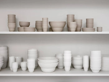 Two white shelves display neatly stacked Vipp Vipp203 Tea Cups, Set of 2 by Vipp, in neutral beige and white tones, organized by size and shape against a plain background.