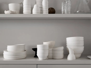 Stacks of premium porcelain dishes and Vipp Vipp218 Bowl, Large by Vipp are neatly arranged on a kitchen counter and shelves, alongside cups, glassware, and a white pitcher against a light gray wall.