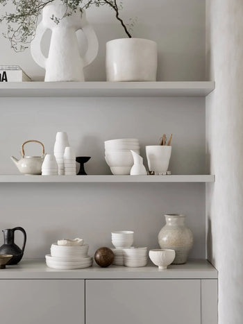 Minimalist shelves display premium porcelain bowls, cups, and vases; decorative pottery; a large Vipp Vipp218 Bowl by Vipp; a teapot; small plant; and a sculptural white figure against a light neutral wall.