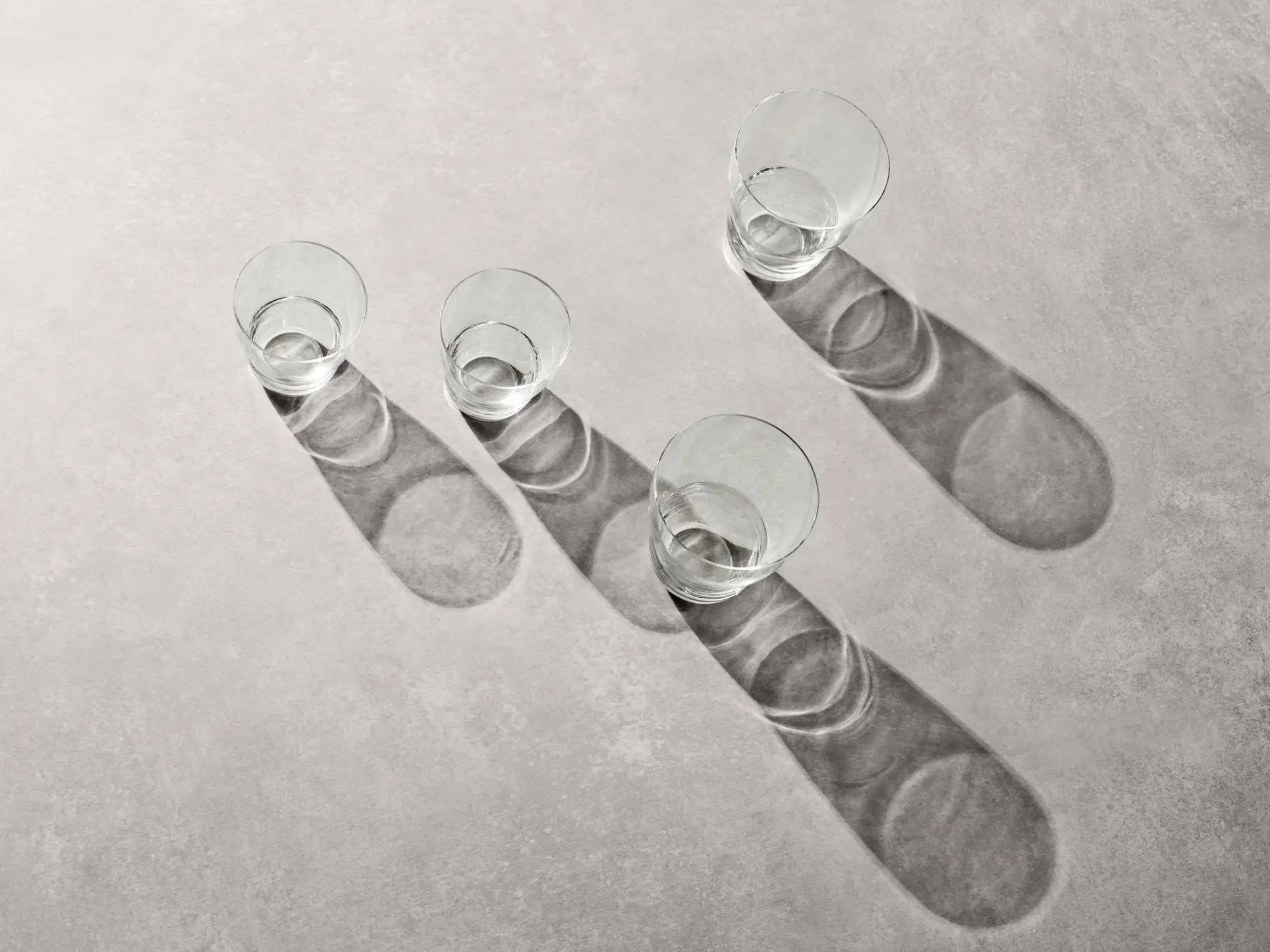Two empty, mouth-blown Vipp Vipp242 Glasses 11oz by Vipp are placed in a diagonal line on a light gray surface, each casting a long shadow to the right from bright, angled lighting.