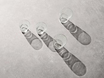 Two empty, mouth-blown Vipp Vipp242 Glasses 11oz by Vipp are placed in a diagonal line on a light gray surface, each casting a long shadow to the right from bright, angled lighting.