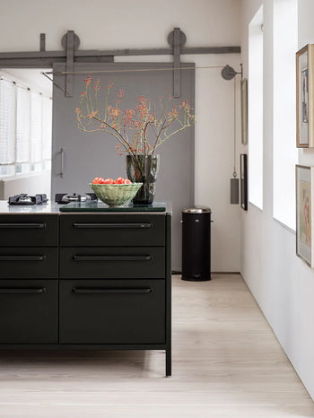 A modern kitchen features a sleek dark island, the Vipp Vipp248 Vase by Vipp with branches, and a bowl of tomatoes. Light wood floors, large windows, and minimalist decor enhance the bright, airy atmosphere.