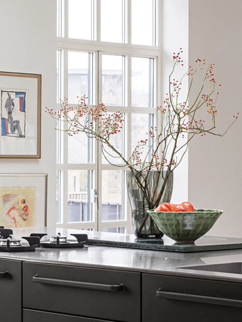 A modern kitchen with a dark countertop features the Vipp Vipp248 Vase by Vipp, filled with branches and red berries, alongside a green bowl of tomatoes and framed artwork below tall windows streaming in natural light.