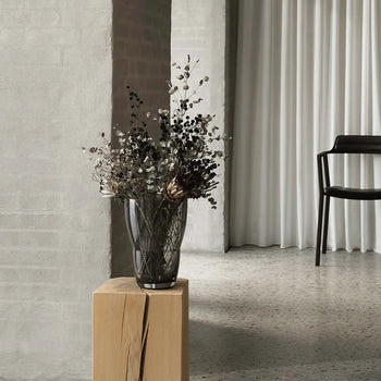 The Vipp Vipp248 Vase by Vipp, displayed with dried flowers on a wooden pedestal, enhances a minimalist room with gray brick walls, terrazzo floor, white curtains, and a black chair in the background.
