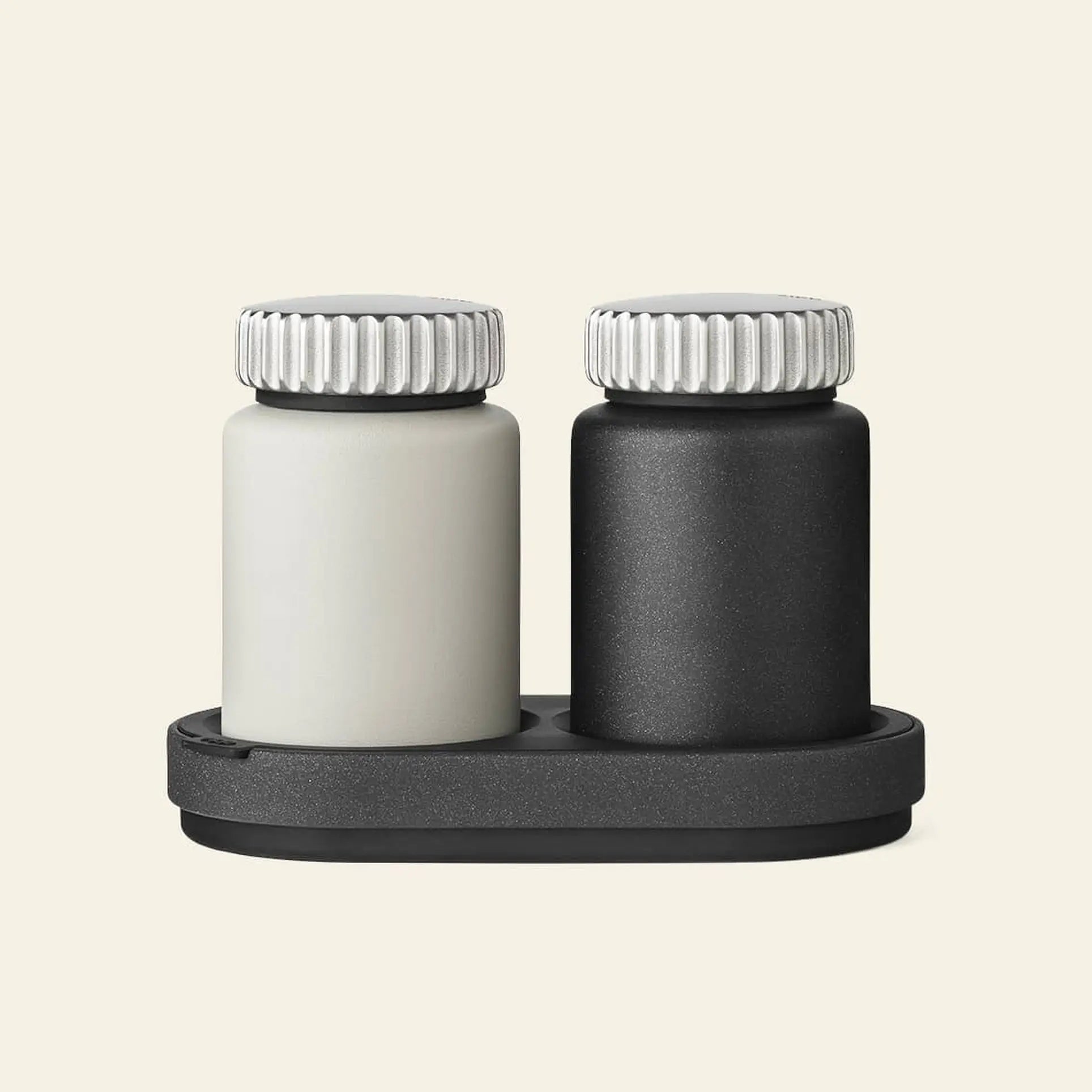 Two small Vipp Vipp263 Salt & Pepper Mills—one white, one black—featuring ribbed silver lids, rest side by side on a dark tray, bringing modern elegance to any kitchen against a plain beige background.