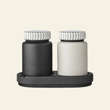 Two small Vipp Vipp263 Salt & Pepper Mills, one black and one white with ribbed silver lids, rest side by side on a matching black tray—a stylish accent for any kitchen, set against a plain beige background.