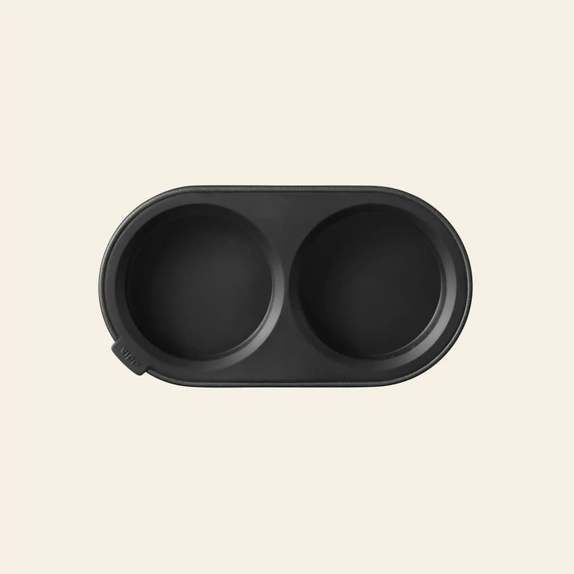 A black, oval silicone pet bowl with two round compartments sits on a beige background—an ideal match for the Vipp Vipp263 Salt & Pepper Mill by Vipp in any culinary space.