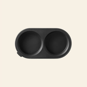 A black, oval silicone pet bowl with two round compartments sits on a beige background—an ideal match for the Vipp Vipp263 Salt & Pepper Mill by Vipp in any culinary space.