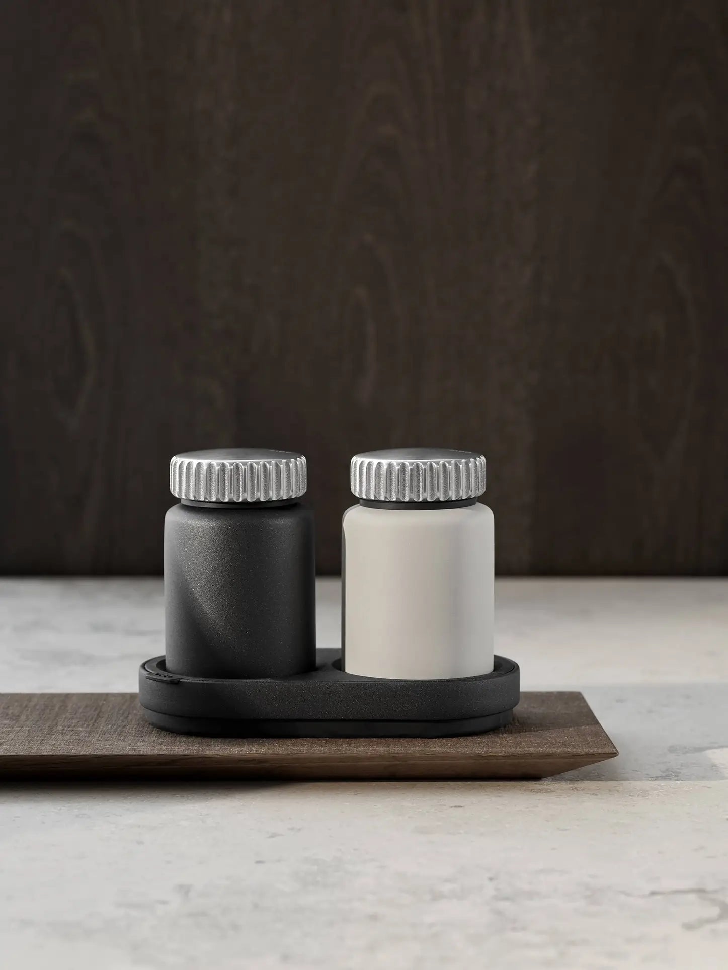 Two Vipp Vipp263 Salt & Pepper Mills—one black, one white—by Vipp with silver lids sit side by side on a black oval tray atop a wooden board, bringing modern style to any kitchen against a dark wood background.