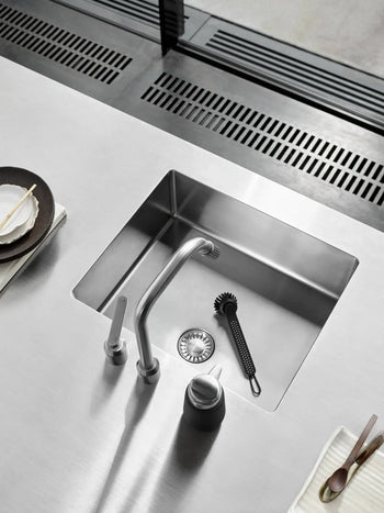 A modern stainless steel kitchen sink features a sleek faucet, a sink strainer, and the Vipp Vipp280 Dishwashing Brush by Vipp with a replaceable head. Plates, cutlery, and napkins are neatly arranged on either side for easy cleaning.