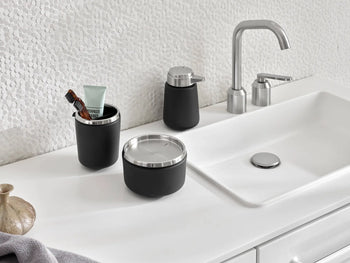 A modern bathroom countertop features a white sink, silver faucet, and black Vipp accessories: a soap dispenser, lidded container, and the Vipp Vipp7 Toothbrush Holder with a detachable rubber inner section. Textured white wall in back.