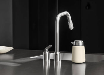 A modern stainless steel kitchen faucet with a matching Vipp Vipp9 Dispenser and beige container sit on a sleek, dark countertop in a minimalist kitchen, showcasing elegant accessory choices from Vipp.