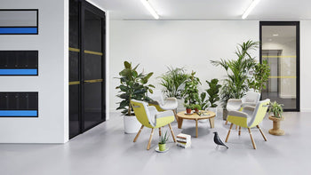 A modern office lounge with green chairs around a wooden table, potted plants, and the Vitra Eames House Bird by Vitra on a light grey floor, creating a bright and welcoming atmosphere inspired by Charles and Ray Eames.
