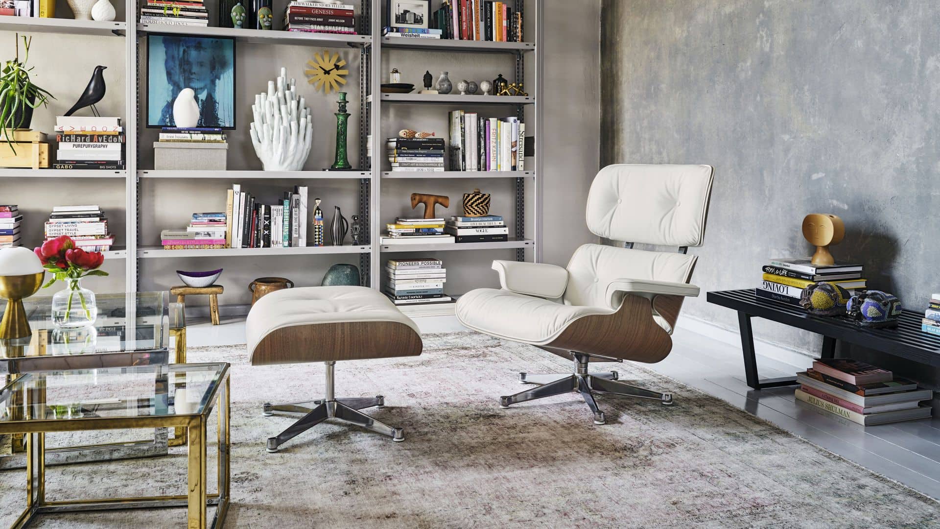 A modern living room featuring a white Vitra Eames lounge chair and ottoman, Vitra Eames House Bird, glass coffee table, shelves with decor, textured gray wall, and a right-side bench displaying decorative items.