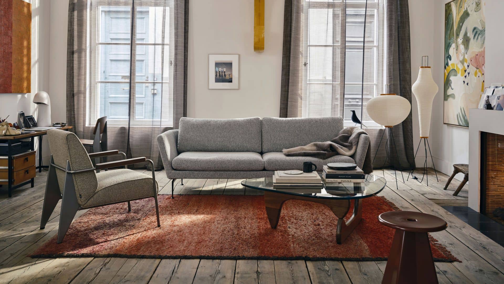A cozy living room with a gray sofa, two armchairs, and a glass coffee table on a red rug showcases stylish decor. Large windows with sheer curtains and the Vitra Eames House Bird by Vitra add charm to this warm, inviting space.