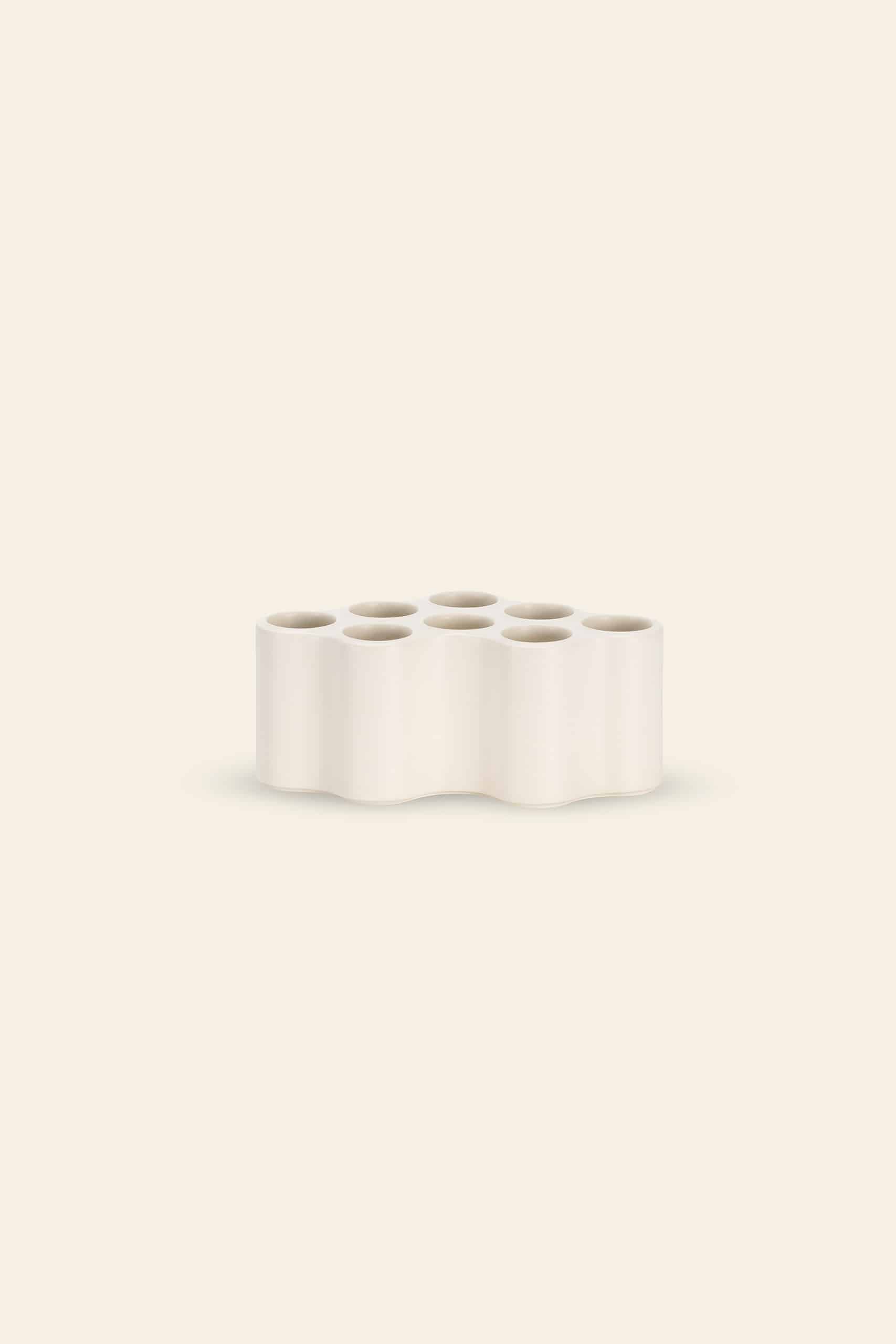 A minimalist, off-white handcrafted ceramic Vitra Nuage Ceramique, Small by Vitra featuring a wavy rectangular shape with six circular openings on top, set against a plain beige background and inspired by ikebana.