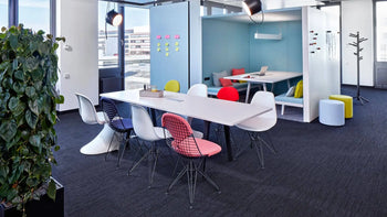 Modern office space with a long white Vitra Toolbox RE table by Vitra, colorful chairs, cubicle area with couches and workstations in the background, large windows, lush plants, and bright lighting.