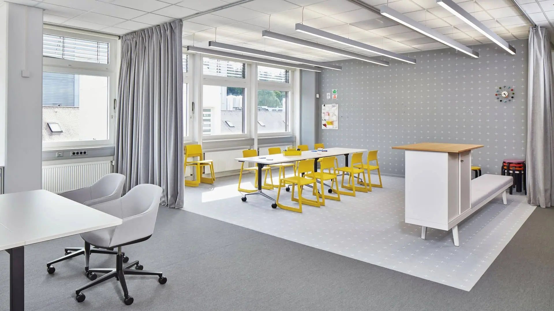 A bright, modern classroom with large windows, yellow chairs, and white tables features the Vitra Wall Clock - Ball Clock by Vitra, its colorful spheres adding a contemporary accent to the clean gray and white decor filled with natural light.